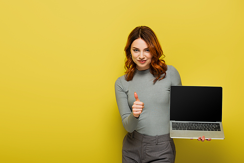 happy woman with curly hair holding laptop with blank screen while showing thumb up on yellow