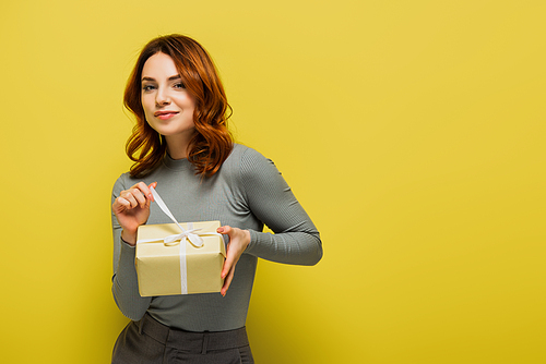 happy young woman pulling ribbon on present and smiling on yellow