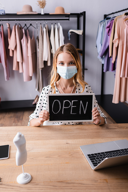 showroom owner in medical mask, holding board with open lettering while sitting at workplace