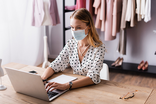 showroom owner in safety mask, typing on laptop at workplace