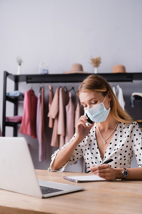 businesswoman in medical mask, talking on smartphone near laptop in showroom
