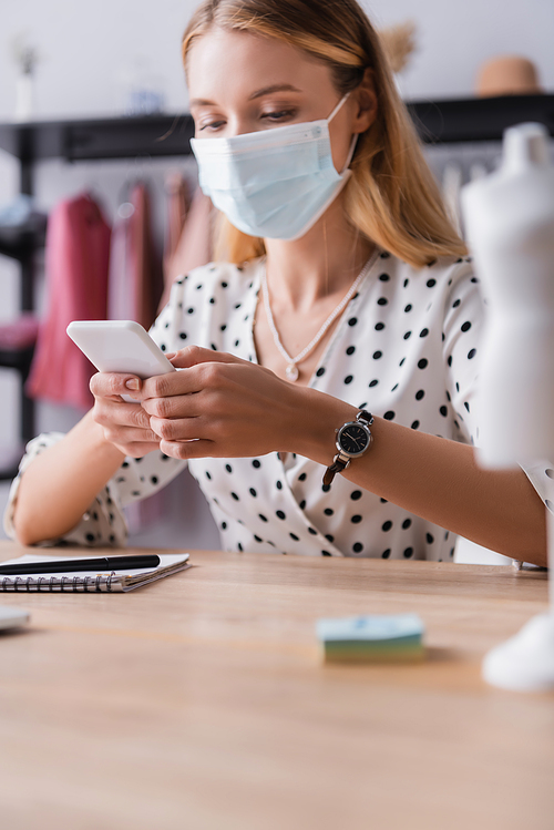 showroom owner in protective mask, messaging on smartphone at workplace on blurred foreground
