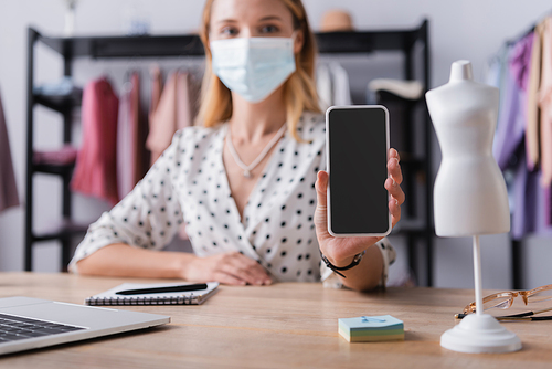 businesswoman in medical mask, showing cellphone with blank screen while working in showroom on blurred background