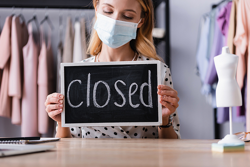 showroom owner in medical mask, holding board with closed lettering on blurred foreground