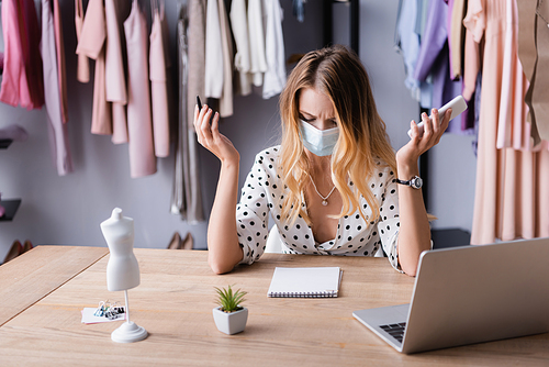 frowning businesswoman in medical mask, holding smartphone and pen near blank notebook in showroom
