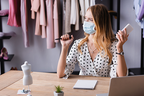thoughtful proprietor in medical mask, holding smartphone and pen while working in showroom