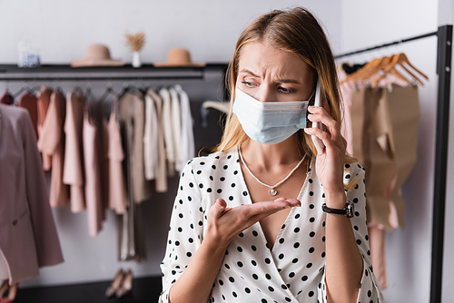 upset businesswoman in medical mask pointing with hand while talking on smartphone in showroom