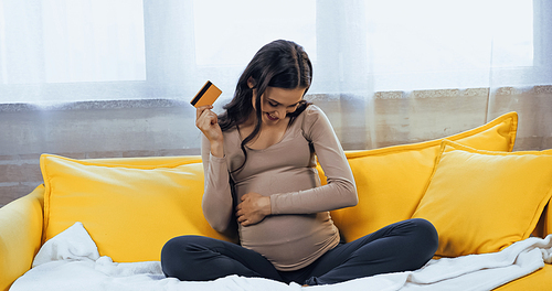 Smiling pregnant woman with credit card looking at belly on couch