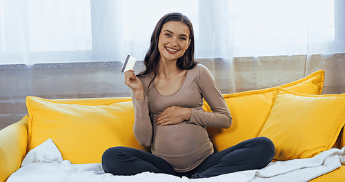 Cheerful pregnant woman holding credit card on couch