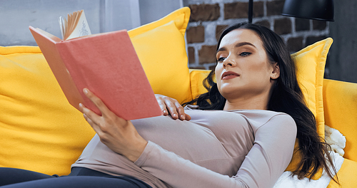 Young pregnant woman reading book while lying on couch