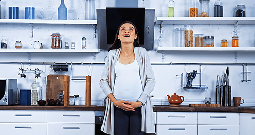 Excited pregnant woman embracing belly in kitchen