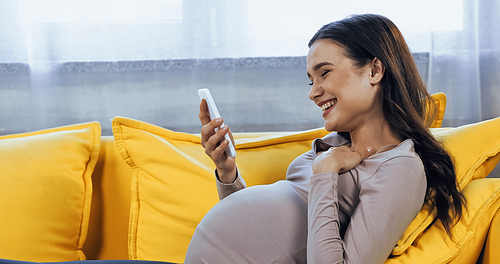 Smiling pregnant woman holding smartphone on bright yellow couch
