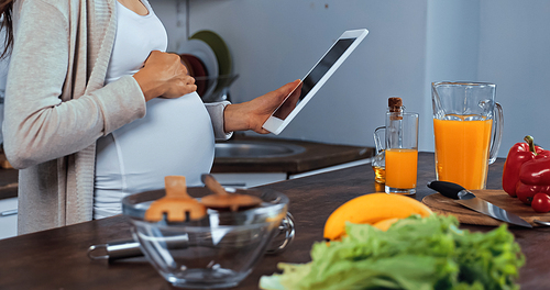 Cropped view of pregnant woman holding digital tablet near vegetables and orange juice in kitchen