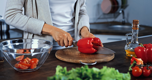 Cropped view of woman cutting bell pepper on cutting board near oil and vegetables on blurred foreground