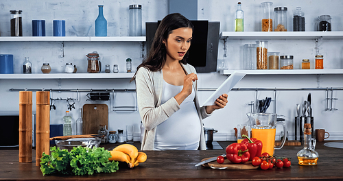 Pregnant woman using digital tablet near food on kitchen table