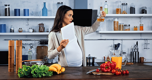 Pregnant woman with digital tablet looking at oil near food on kitchen table