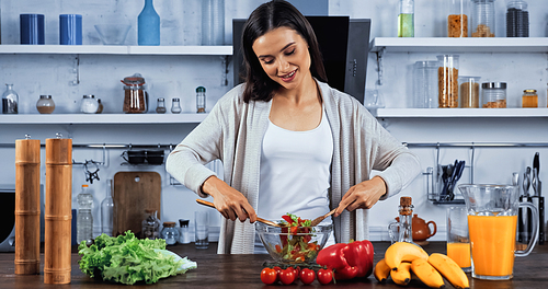 Smiling woman mixing fresh salad near orange juice on kitchen table