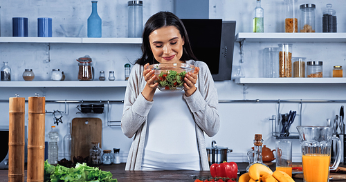 Smiling woman holding bowl with fresh salad near vegetables and orange juice