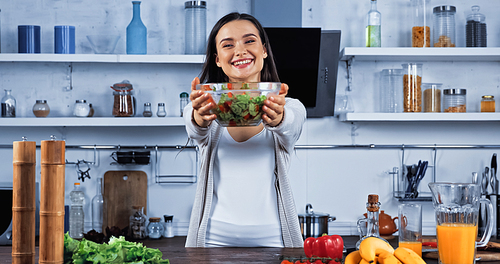 Smiling woman showing bowl of salad near fresh food on kitchen table