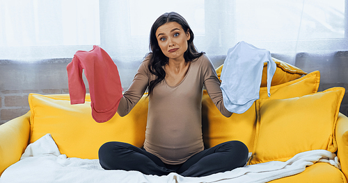 Confused pregnant woman holding baby clothes on couch