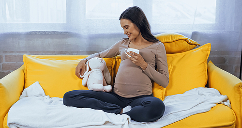 Pregnant woman with cup looking at soft toy while sitting on yellow couch