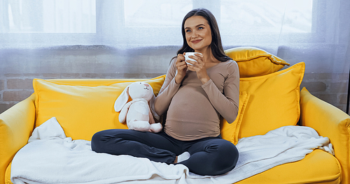Dreamy pregnant woman with cup sitting near soft toy