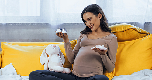 Smiling pregnant woman looking at baby booties near soft toy on couch