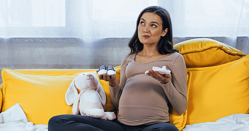 Pensive pregnant woman holding baby booties near soft toy