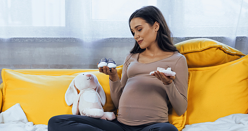 Pregnant woman holding baby booties near soft toy at home