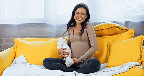 Pregnant brunette woman smiling at camera and holding soft toy on couch