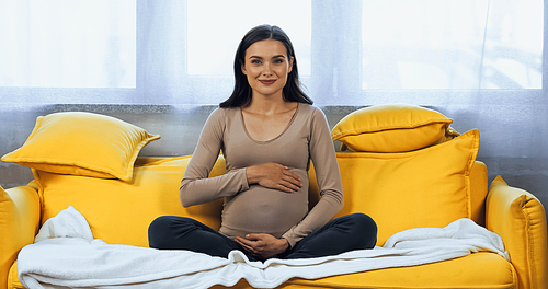 Pregnant woman hugging belly and smiling at camera on sofa