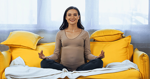 Cheerful pregnant woman sitting in lotus pose on couch