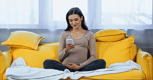 Pregnant brunette woman smiling and using smartphone on couch