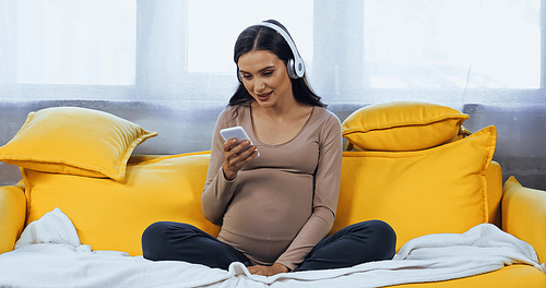 Smiling pregnant woman in headphones using smartphone in living room