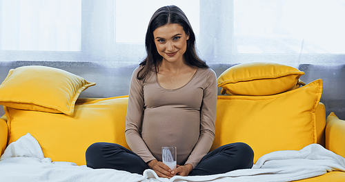 Pregnant woman smiling at camera while holding glass of milk at home