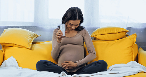 Pregnant woman holding glass of fresh milk and looking at belly on sofa