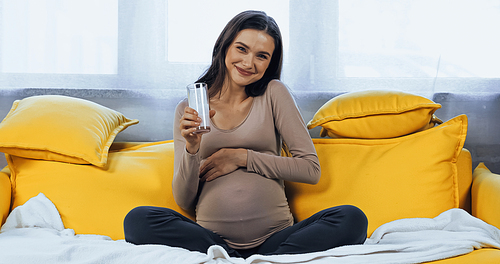 Happy pregnant woman holding glass of milk of yellow couch at home