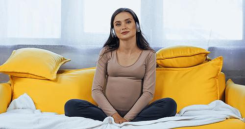 Young pregnant woman listening music in headphones on couch