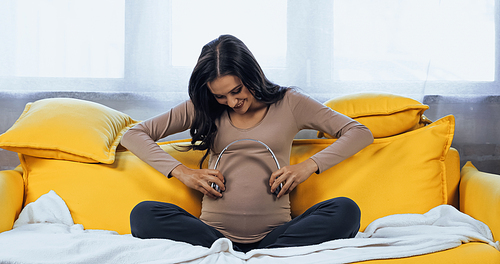 Happy pregnant woman holding headphones near belly while sitting on couch