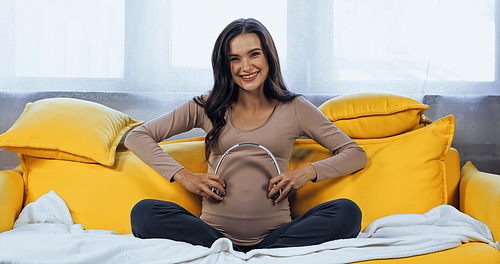 Cheerful pregnant woman holding headphones near belly