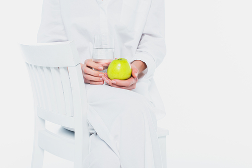Cropped view of woman in white clothes holding apple and glass of water on chair isolated on white
