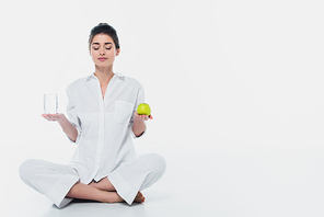 Brunette woman with glass of water and apple sitting in yoga pose on white background