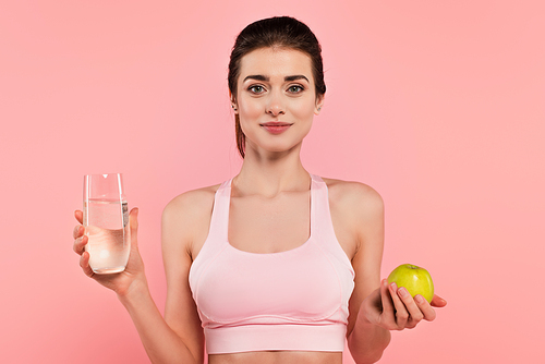 Young sportswoman holding green apple and glass of water isolated on pink