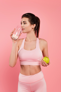 Brunette sportswoman drinking water and holding apple isolated on pink