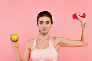 Young sportswoman holding dumbbell and apple isolated on pink