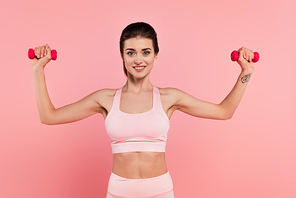 Cheerful sportswoman training with dumbbells isolated on pink