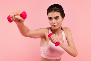 Pretty sportswoman training with dumbbells on blurred foreground isolated on pink