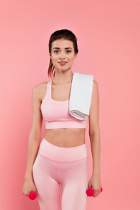 Smiling sportswoman with towel holding dumbbells isolated on pink