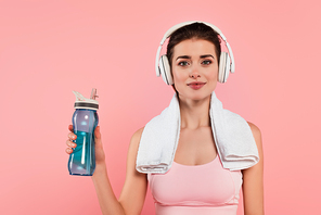 Brunette sportswoman in headphones and towel holding sports bottle isolated on pink