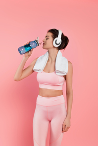 Sportswoman with headphones and towel drinking water isolated on pink